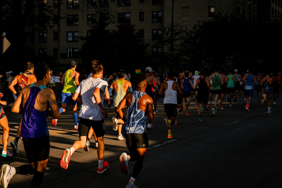 An action shot of people running the Chicago Marathon. Runners are wearing colorful running clothes, their backs are turned to the camera. The sunlight shines down on half of the runners while the others run in shadow.