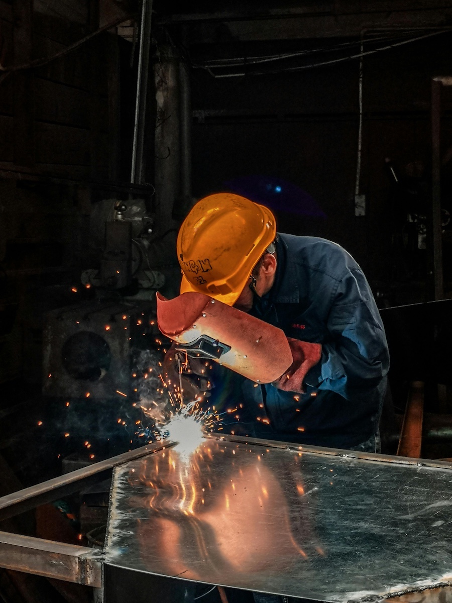 Person wearing orange hard hat and protective welder's mask cutting large metal piece, sparks flying from cutting tool