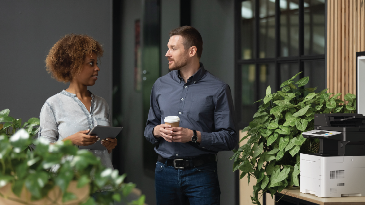 media-image-1178×663-unlimited-plan-kyocera Two office employees in a contemporary office environment standing near a Kyocera copier on a table, surrounded by greenery from potted plants